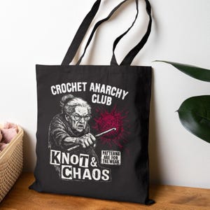May include: Black tote bag with the words "CROCHET ANARCHY CLUB" and "KNOT & CHAOS" printed in white. The graphic features an illustration of an elderly woman crocheting with a red yarn ball and the text "PATTERNS ARE FOR THE WEAK".
