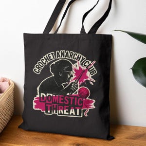 May include: Black tote bag featuring a graphic of a woman crocheting with the text "CROCHET ANARCHY CLUB" above and "DOMESTIC THREAT" below. The design includes pink and white accents.