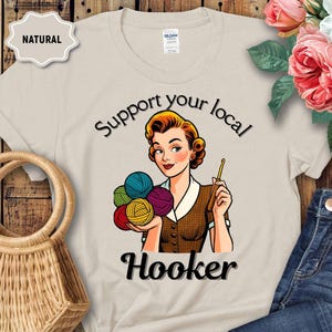 May include: A cream-coloured t-shirt featuring a vintage-style illustration of a woman with yarn and a crochet hook. The text says "Support your local Hooker". The shirt has a round neck and short sleeves. The word "Natural" is printed on a beige label.