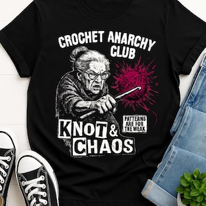 May include: Black t-shirt with a graphic featuring an elderly woman crocheting, the text "CROCHET ANARCHY CLUB," and "KNOT & CHAOS." A red yarn ball and the phrase "PATTERNS ARE FOR THE WEAK" are also included.