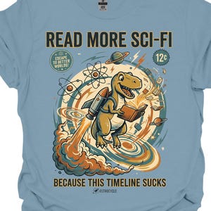 May include: Stone blue t-shirt with a graphic featuring a cartoon dinosaur riding a rocket and reading a book. Text on the shirt reads "READ MORE SCI-FI BECAUSE THIS TIMELINE SUCKS". The design includes planets, atoms, and the words "ESCAPE TO BETTER WORLDS!"