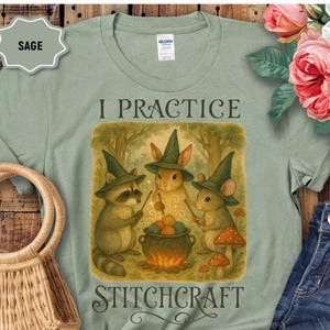 May include: Sage green t-shirt with the words "I Practice Stitchcraft". The graphic features a raccoon, rabbit, and mouse wearing witch hats, gathered around a cauldron with yarn. The design includes mushrooms and a forest backdrop.