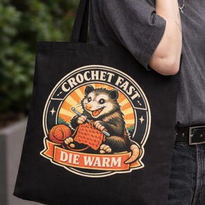 May include: Black tote bag featuring a cartoon opossum crocheting with the text "CROCHET FAST DIE WARM". The opossum is brown and gray, with orange yarn and a crochet hook. The background has a sunset design.