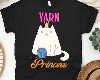 May include: Black t-shirt featuring a white cat wearing a crown and holding a blue ball of yarn. The text "YARN" is above the cat in pink, and "Princess" is below in a gold script font. A fun design for yarn lovers.