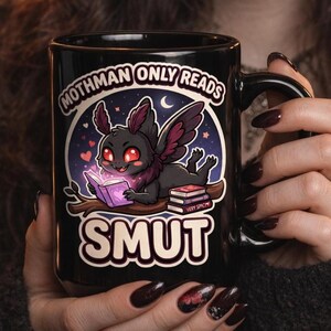 May include: Black ceramic mug featuring a cartoon Mothman reading a book with the text "Mothman Only Reads Smut." The design includes a Mothman illustration with red eyes and purple wings, set against a starry background.