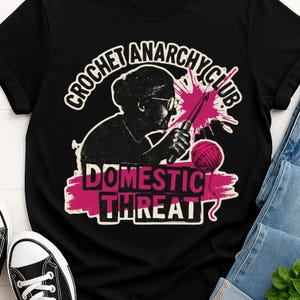 May include: Black t-shirt featuring a graphic of a woman crocheting with the text "CROCHET ANARCHY CLUB" above and "DOMESTIC THREAT" below. The design includes a pink yarn ball and a splash of pink. The shirt is paired with blue jeans and black sneakers.