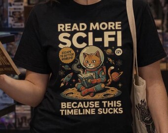 May include: A black t-shirt featuring a graphic of an orange cat in a space suit sitting on the moon reading a book. The text reads: READ MORE SCI-FI, ESCAPE TO BETTER WORLDS, 12c, and BECAUSE THIS TIMELINE SUCKS. The design includes planets and rockets.