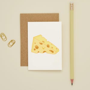 Könnte beinhalten: Eine Grußkarte mit einer Cartoon-Illustration eines Stücks Schweizer Käse mit einem lächelnden Gesicht. Die Karte befindet sich auf weißem Hintergrund, mit einem braunen Umschlag dahinter. Ein gelber Bleistift und zwei goldene Büroklammern sind ebenfalls im Bild.