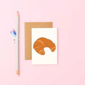 Könnte beinhalten: Eine Grußkarte mit einer Cartoon-Illustration eines lächelnden Croissants. Die Karte befindet sich auf weißem Hintergrund, mit einem braunen Umschlag dahinter. Ein hellgrüner Bleistift und Briefmarken befinden sich auf rosa Hintergrund.