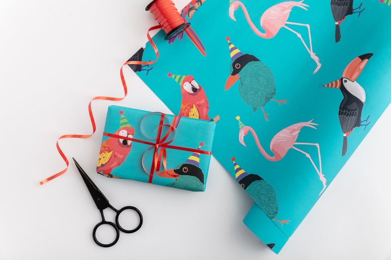 May include: Turquoise blue wrapping paper with a repeating pattern of pink flamingos, parrots, toucans, and black birds wearing party hats. The paper is rolled up and partially unrolled, revealing the pattern. A red ribbon and a pair of black scissors are also in the image.