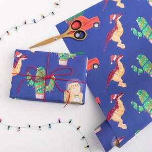 May include: A blue wrapping paper with a repeating pattern of dinosaurs wearing Santa hats, cacti with Christmas lights, and a robot with a snow globe. A red toy truck is also part of the pattern. A gift box wrapped in the same paper is tied with red string.
