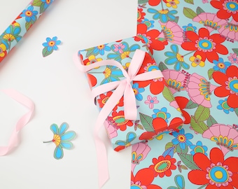 May include: A gift wrapped in blue and white floral patterned paper with a pink ribbon. The gift is sitting on a white surface next to a roll of wrapping paper with a similar pattern.