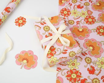 May include: A pink gift box with a white ribbon is wrapped in floral patterned paper. The paper features a repeating pattern of pink, orange, and yellow flowers on a light pink background.