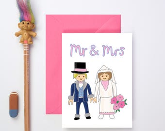 Pode incluir: Um cartão branco com o texto "Mr & Mrs" em letras roxas. O cartão apresenta uma ilustração de desenho animado de uma noiva e um noivo com roupas azuis e brancas.