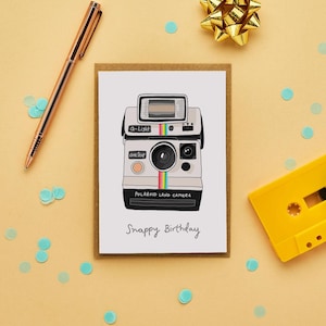 Puede incluir: Tarjeta de cumpleaños con una ilustración de cámara Polaroid retro. La cámara es blanca con una franja colorida y las palabras "Polaroid Land Camera" y "Snappy Birthday". También hay un bolígrafo dorado, confeti y una cinta de cassette amarilla.