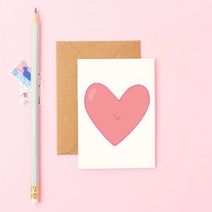 Puede incluir: Una tarjeta de felicitación con una ilustración de corazón rosa sonriente sobre fondo blanco. Un sobre marrón y un lápiz verde claro están junto a la tarjeta. El fondo es de color rosa liso. Hay dos sellos cerca del lápiz.