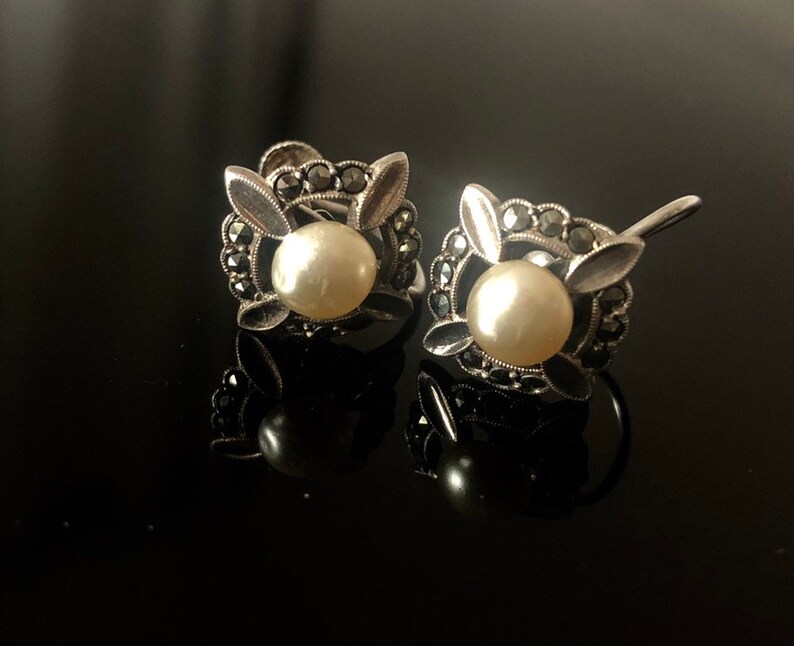 May include: A pair of silver earrings with a pearl and marcasite stones in a floral design.