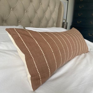 Pu&ograve; includere: Un cuscino lombare marrone con strisce verticali bianche e punti bianchi. Il cuscino &egrave; su un letto bianco con una testata imbottita.