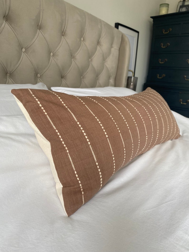 Pu&ograve; includere: Un cuscino lombare marrone con strisce verticali bianche e punti bianchi. Il cuscino &egrave; su un letto bianco con una testata imbottita.