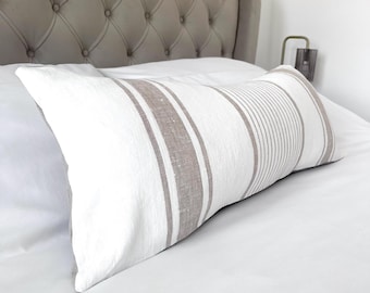 May include: A rectangular white pillow with vertical brown stripes and lines. The pillow is placed on a white bed with a gray button-tufted headboard in the background. The pillow is a decorative element for the bed.
