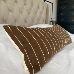 Peut inclure: Un oreiller rectangulaire marron avec des motifs de points et de rayures blancs. L'oreiller est posé sur un lit blanc, avec une tête de lit capitonnée en arrière-plan. L'arrière de l'oreiller est de couleur crème.