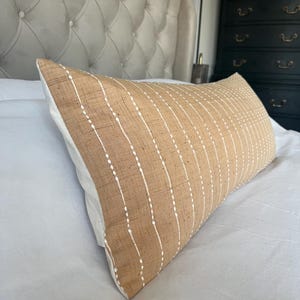 Puede incluir: Una almohada de tiro a rayas beige y blanca con un patrón de puntos blancos. La almohada está sobre una cama blanca con una cabecera capitoné.