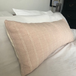 Puede incluir: Una almohada lumbar de color rosa claro con rayas blancas bordadas. La almohada está sobre una cama blanca.
