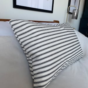 May include: A white and black striped pillow with a subtle texture. The pillow is lying on a white bed.