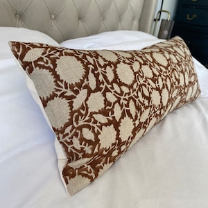 May include: A rectangular cushion with a brown floral design on a beige fabric. The cushion is placed on a white bed, with a buttoned headboard in the background. The floral pattern consists of repeating stylized flowers and leaves.