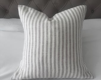 May include: A square pillow with a gray and white striped pattern. The pillow is resting on a white surface, with a gray, tufted headboard in the background. The pillow's fabric has a textured appearance.