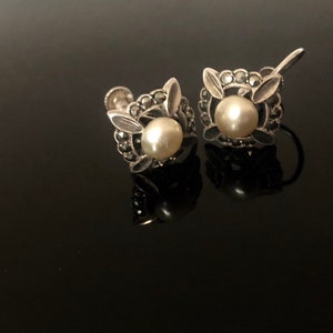 May include: A pair of silver earrings with a pearl and marcasite design. The earrings are in the shape of a flower with a pearl in the center and marcasite stones around the pearl.