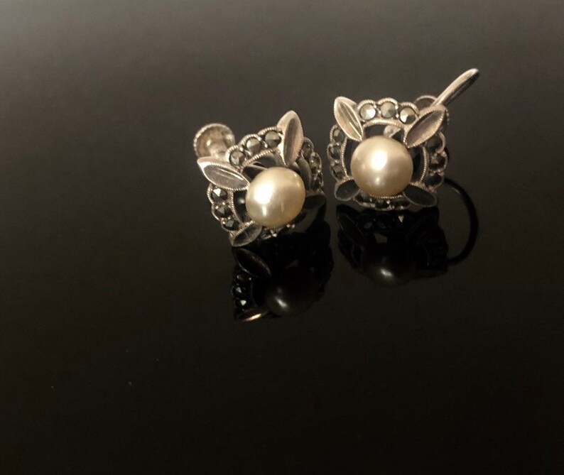 May include: A pair of silver earrings with a pearl and marcasite design. The earrings are in the shape of a flower with a pearl in the center and marcasite stones around the pearl.
