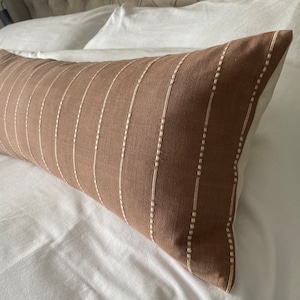 Pu&ograve; includere: Un cuscino lombare marrone con strisce bianche ricamate. Il cuscino &egrave; su un letto bianco.
