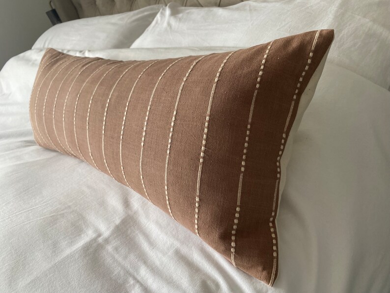 Pu&ograve; includere: Un cuscino lombare marrone con strisce bianche ricamate. Il cuscino &egrave; su un letto bianco.