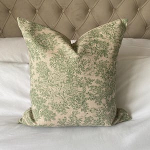 May include: A green and white patterned cushion with a floral design. The cushion is on a white bed with a brown headboard.