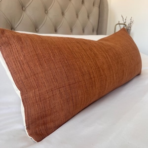 May include: A long, rectangular cushion with a textured, rust-coloured fabric cover. The cushion has white edges and rests on a white bed. The headboard in the background is a light grey colour.
