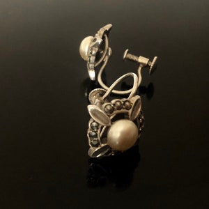 May include: A pair of silver clip-on earrings with a pearl and marcasite design. The earrings have a floral motif with a single pearl in the center of each earring.