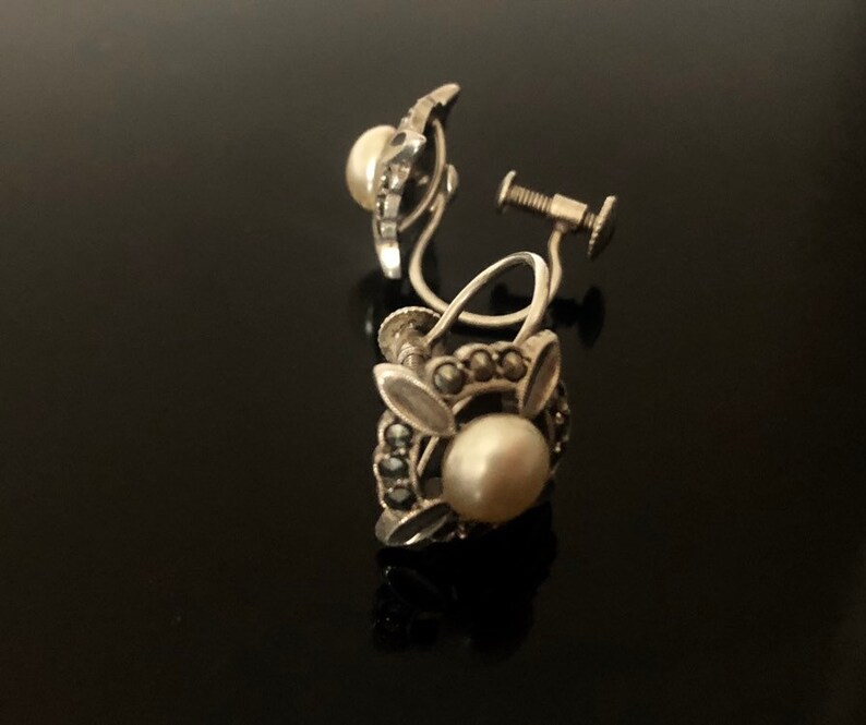 May include: A pair of silver clip-on earrings with a pearl and marcasite design. The earrings have a floral motif with a single pearl in the center of each earring.