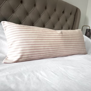 Pode incluir: Uma almofada retangular com fundo creme e finas listras vermelhas horizontais. A almofada está colocada sobre uma cama branca com cabeceira almofadada. A almofada mede aproximadamente 91 cm de comprimento.