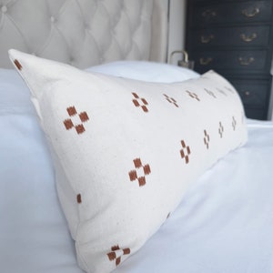 May include: A white lumbar pillow with a brown geometric pattern. The pillow is lying on a white bed with a tufted headboard.