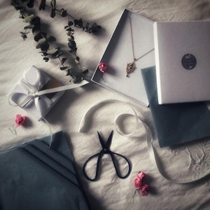 May include: A white gift box with a white ribbon and bow, a blue velvet box with a gold eye pendant necklace, a white gift box with the text "Sweet Dreams" and a pair of black scissors on a white surface.