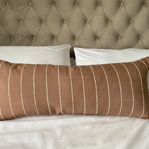May include: A brown lumbar pillow with white vertical stripes and small white dots. The pillow is on a white bed with a tufted headboard.