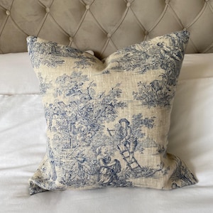 Op de afbeelding: Een blauw en wit toile-patroon kussen met een linnen textuur. Het kussen heeft een landelijke scène met figuren en bomen.