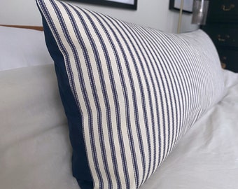 May include: A long, rectangular pillow with a white and navy blue striped pattern. The pillow has a navy blue border and is lying on a white bed.
