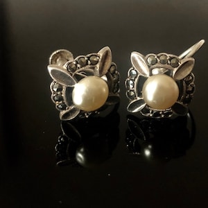 May include: A pair of silver earrings with a square design. Each earring features a small pearl surrounded by black marcasite stones.