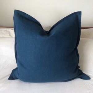 May include: A deep blue square cushion with a simple design, made of a soft, textured fabric. The cushion is lying on a white bedspread.