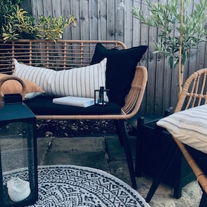 Pu&ograve; includere: Un divano in vimini per patio con cuscini neri e un cuscino a righe. Una lanterna nera con una candela bianca si trova su un tappeto a motivi neri e bianchi davanti al divano.