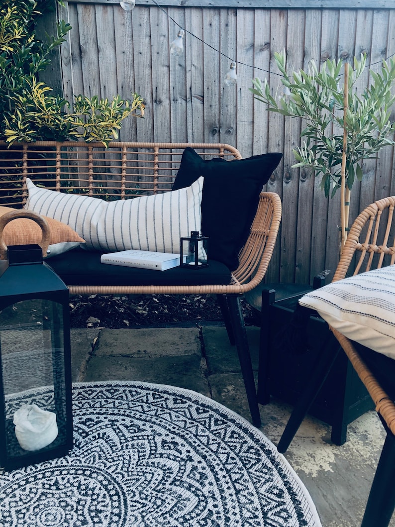Pu&ograve; includere: Un divano in vimini per patio con cuscini neri e un cuscino a righe. Una lanterna nera con una candela bianca si trova su un tappeto a motivi neri e bianchi davanti al divano.