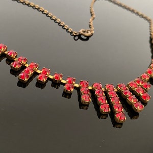 Pode incluir: Um colar de corrente de ouro com um design em cascata de barras de strass vermelhas. O colar está sobre uma superfície preta reflexiva.