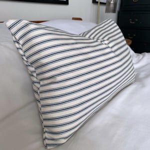 May include: A white and blue striped pillow with a button detail. The pillow is on a white bed with a wooden headboard.